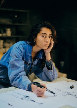 Porträt der Künstlerin Matilde Romagnoli in blauem Arbeitsanzug, am Tisch lehnend bei der Skizzenarbeit im Atelier.